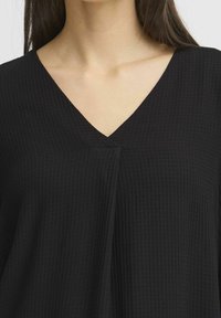 Top noir en v avec un tissu texturé, présentant un détail de pli discret au niveau du col et une coupe décontractée pour le confort.