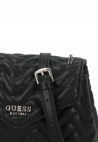 Borsa a tracolla nera trapuntata con superficie testurizzata, caratterizzata da un logo argentato "GUESS EST 1981" e una tracolla simile a una cintura con fibbia metallica.