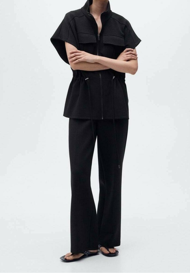 Personne portant une veste sans manches noire zippée avec des cordons, un pantalon noir à jambes larges et des sandales noires, debout, les bras croisés.