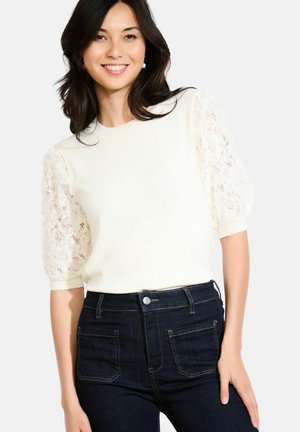 Pull crème léger avec des manches bouffantes en dentelle texturée, col rond et poignets côtelés, associé à un jean taille haute en denim foncé.