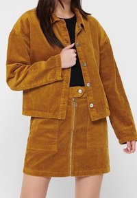 Giacca in corduroy senape con chiusura a pulsante e due tasche anteriori, abbinata a una mini gonna coordinata con zip frontale e tasche.