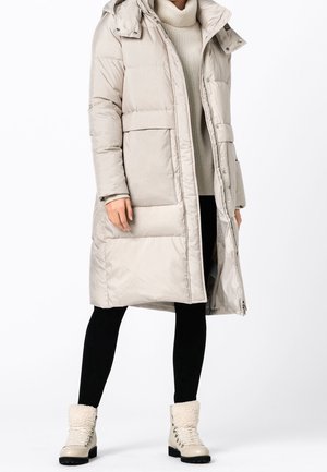 Personne portant un long manteau doudoune beige, un pull à col roulé crème, des leggings noirs et des bottes d'hiver beiges doublées de fourrure.
