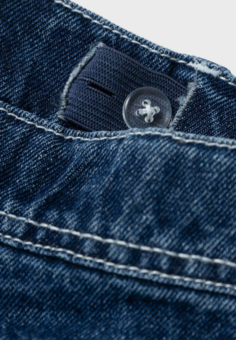 Donkerblauwe denimstof met een plastic knoop en contrasterende witte stiksels, met een labelgedeelte zichtbaar nabij de knoop.