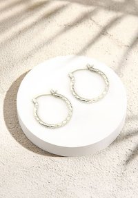 Silverfärgade hoopörhängen med en flätad textur och geometriskt mönster. Cirkelformade med ett säkert spänne, placerade på en rund vit bas.