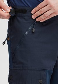 Tumši zilas cargo bikses ar velcro un rāvējslēdzēju aizdari, ar sānu kabatām un izturīgu auduma tekstūru. Regulējama josta komfortam.