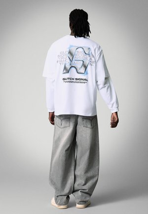 Osoba z dredami ubrana w białą koszulę z długim rękawem z dużym logo stylizowanym na glitch art i napisem na plecach, w połączeniu z luźnymi szarymi jeansami.