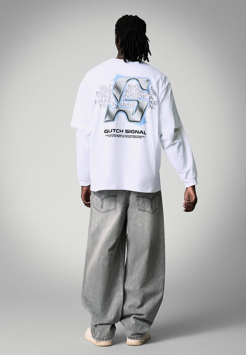 Osoba s dredy, oblečená v bílém tričku s dlouhým rukávem s velkým logem glitch art a textem na zádech, doplněná volnými šedými džínami.