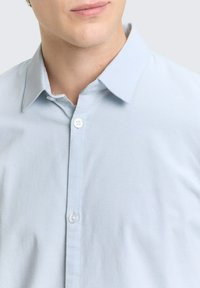 Man in een lichtblauwe overhemd met knoopjes en een kraag, toont het lagere deel van het gezicht en het bovenste deel van de torso.