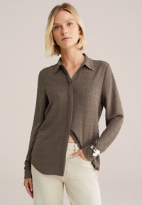 Le modèle pose avec assurance dans une chemise en olive texturé, associée à un pantalon clair, devant un fond neutre, mettant en valeur un style moderne.