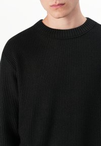 Pull en tricot noir avec un col rond et un motif texturé. Le tissu semble doux avec une finition côtelée et des accents minimaux.