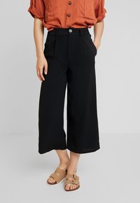 Pantalons larges noirs à taille haute, avec des plis et des poches latérales, associés à des sandales à boucle marron clair et une chemise orange à boutons.