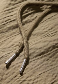 Cuerda trenzada color caqui con puntas de metal plateado sobre un fondo de tela texturizada en tono beige, que presenta detalles de costura sutiles.