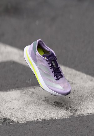 Chaussure de course violet clair et grise avec des lacets noirs, en suspension au-dessus d'une surface en bitume grise avec des marquages blancs.