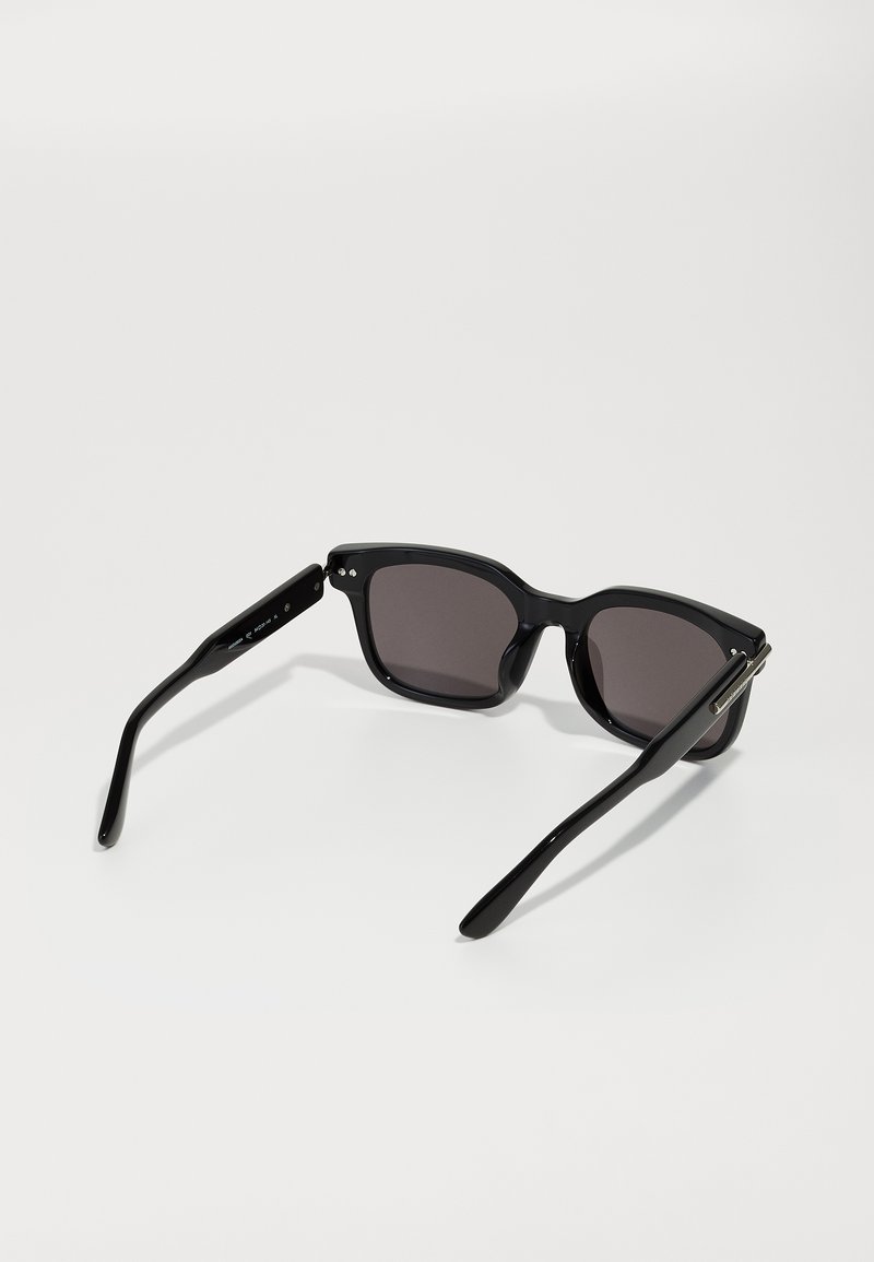 Lunettes de soleil noires avec des montures rectangulaires, des verres teintés foncés et des branches fines ; finition noire brillante et accents métalliques sur les charnières.