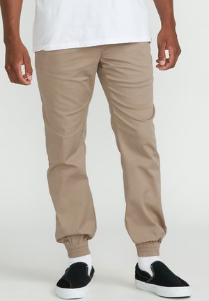 Beige tapered joggerbukser med elastisk talje og manchetter ved anklerne, parret med sorte slip-on sko og hvide crew-sokker.