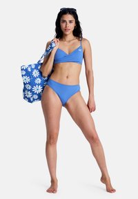 Μπλε σετ μπικίνι με τοπ τύπου wrap και ταιριαστά κάτω μέρη. Κρατώντας μια μπλε τσάντα tote με λευκά ανθισμένα μοτίβα.