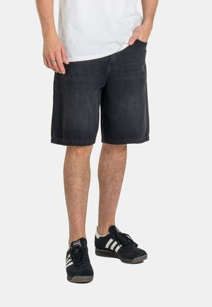 Mann trägt schwarze Jeansshorts, weißes T-Shirt und schwarze Adidas-Sneaker mit weißen Streifen und steht mit einer Hand in der Tasche.