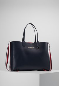 Navy skórzana torba na ramię z podwójnymi uchwytami, akcentami w czerwono-białe paski oraz złotym logo z przodu. Gładka tekstura i przestronny design.