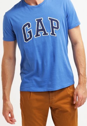 Blå bomulds T-shirt med fede hvide bogstaver, der staver "GAP" i midten. Korteærmer og rund halsudskæring. Matchet med brune bukser.