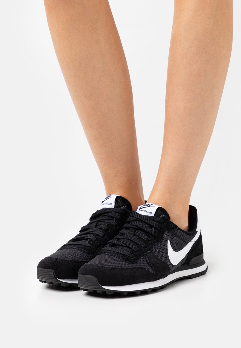 Schwarze Sportschuhe aus Wildleder und Mesh-Material; weißer Nike-Swoosh; schwarze Schnürsenkel; strukturierte schwarze Sohle; flaches Design.