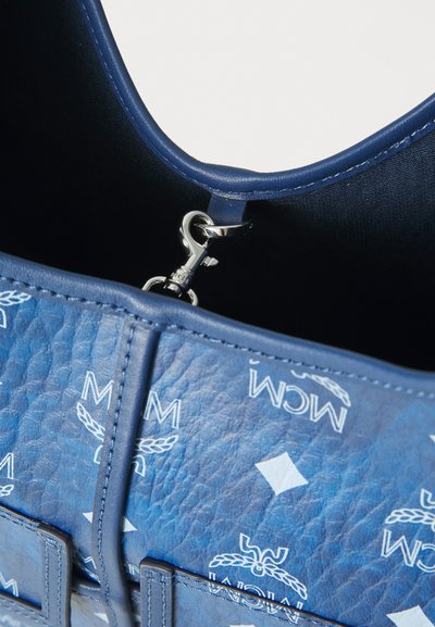 Μπλε υφασμάτινη δερμάτινη τσάντα με εσωτερικό λείο, ασημένιο κλείσιμο και λευκό μοτίβο λογότυπου MCM στην εξωτερική πλευρά.