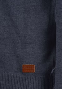 Granatowy sweter dziergany z prążkowanym wykończeniem na dole i przy mankietach, z brązową skórzaną metką z wytłoczonym napisem "BLEND" u dołu. Miękka faktura.