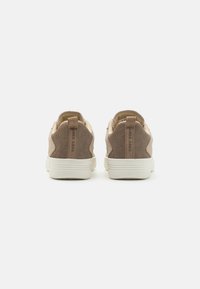 Brune semskede sneakers med en hvit gummisåle, med en teksturert hæl og minimalistisk design. Bakvinduet viser løftestropper for enkel påkledning.