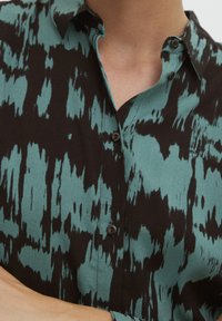 Skjorta med knäppning framtill som har ett svart och teal abstrakt mönster, tillverkad av ett mjukt tyg. Kort krage och svarta knappar förhöjer designen.