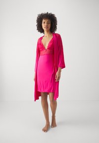 Robe en satin rose avec des bordures en dentelle et un décolleté en V profond, superposée sous une robe assortie. Texture lisse et silhouette ajustée avec un drapé doux.