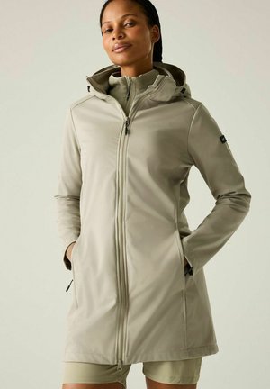 Mujer con una chaqueta larga con cremallera de color beige claro, con capucha, bolsillos y cuello alto, de pie frente a un fondo liso y claro.