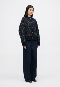 Cardigan marine avec motif argyle dans des couleurs claires, grands boutons et ourlet à côtes ; associé à un pantalon en denim bleu foncé à jambes larges et des chaussures noires.