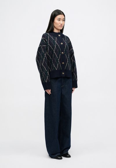 Cárdigan navy con patrón de rombos en colores claros, grandes botones y dobladillo acanalado; combinado con pantalones de mezclilla oscuros de pierna ancha y zapatos negros.