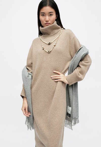 Bež haljina s rolkom i rebrastim manšetama, nošena s svijetlosivim šal. Naglašena slojevitim ogrlicama u zlatnoj boji.