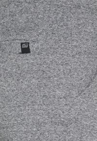Tela texturizada gris con una pequeña etiqueta rectangular en blanco y negro cosida cerca de la apertura del bolsillo.