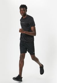 Camiseta atlética negra y pantalones cortos, con un logo reflectante en el pecho, combinados con zapatillas negras con textura. Diseño sencillo y ajustado.