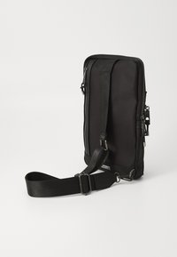 Sort nylon taske med justerbar rem, rektangulær form, sølvfarvet hardware, dobbelt lynlås lukning og minimalistisk design. Textureret overflade.