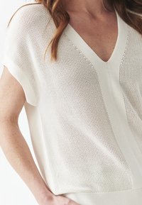 Žena nosící bílý krátký rukáv, volně pletený svetr s výstřihem do V a žebrovaným lemem, odhalující rameno, ruku a dlaň opřenou o pas.