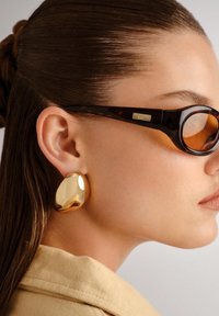 Lunettes de soleil écaille de tortue avec verres foncés et boucles d'oreilles en forme abstraite de ton doré. Le modèle a les cheveux tirés en arrière et une carnation claire.