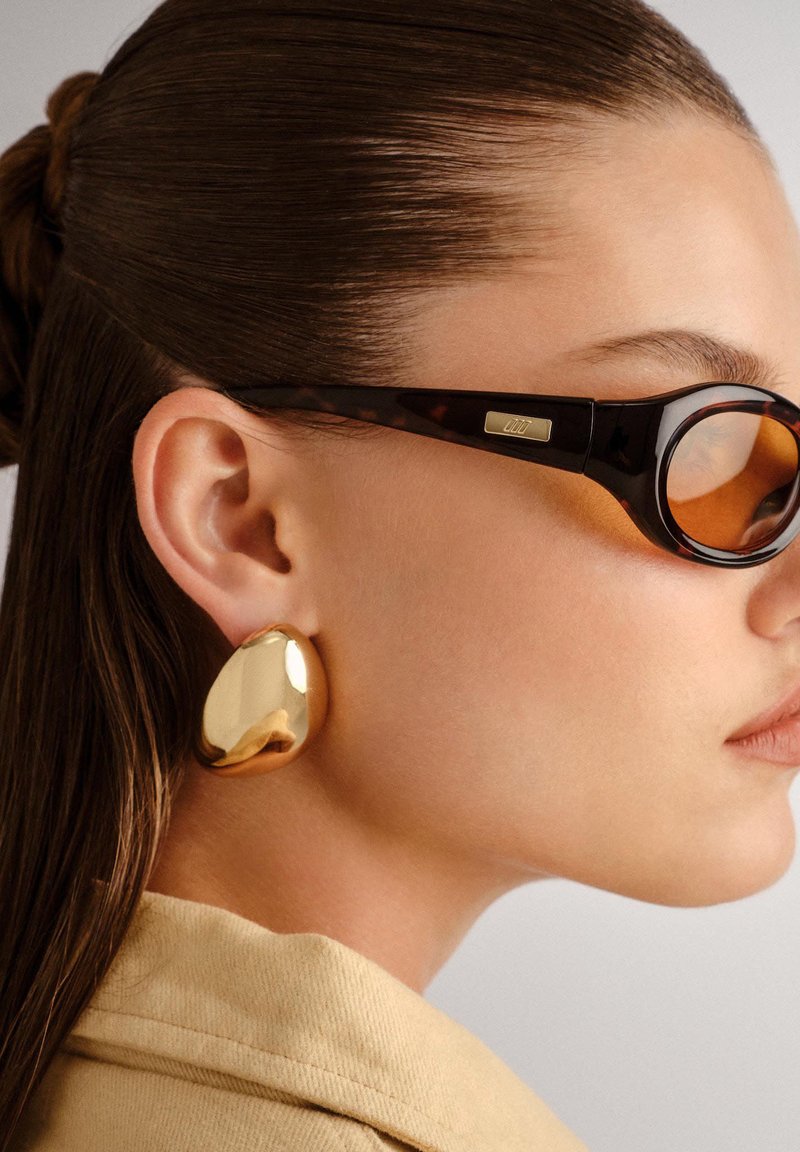 Lunettes de soleil écaille de tortue avec verres foncés et boucles d'oreilles en forme abstraite de ton doré. Le modèle a les cheveux tirés en arrière et une carnation claire.