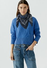 Blauwe gebreide trui met ribbelzoom en zak, gecombineerd met high-waisted vervaagde spijkerbroek. Een marineblauwe gebloemde sjaal is losjes om de hals geknoopt.