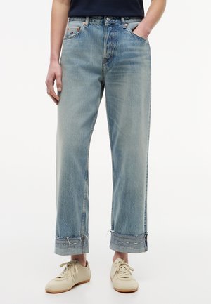 Person iført lyseblå falmede jeans med opsmøgede kanter og beige snøresneakers, stående mod en ensfarvet baggrund.