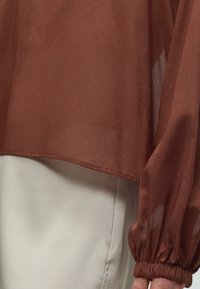 Blouse marron transparente à manches longues avec poignet élastique froncé portée sur un pantalon beige clair, montrant la couture latérale et la main près du poignet.