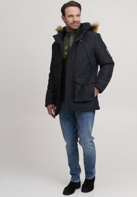 Black insulated parka with fur-lined hood, zip and button closure, two front pockets, and red accents, paired with blue jeans and black boots.