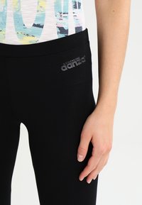 Leggings noirs en matière extensible avec une texture lisse. Présente un logo argenté sur la hanche gauche. Pas de motifs ni d'accents visibles.
