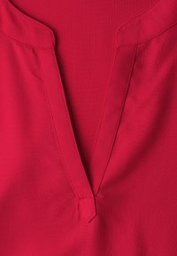 Gros plan sur l'encolure d'un vêtement rouge avec une encochure en V, présentant des coutures visibles et des bords de col pliés.