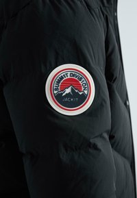 Juodas paminkštintas striukės rankovė su apvaliu ženkleliu, kuriame vaizduojami kalnai, raudona saulė ir tekstas "SUMMIT DIVISION JACK1T DGC."