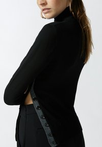 Pull en maille côtelée noir à col roulé avec un côté légèrement ouvert agrémenté de boutons bleu marine. Design ajusté, texture douce.