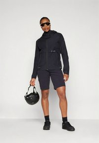 Juoda atspari vandeniui striukė su kapišonu, užtrauktuku ir šoninėmis kišenėmis. Derinama su tamsiais šortais ir laiko juodą šalmą.