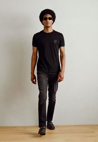 Zwarte t-shirt met een subtiel logo, gecombineerd met donkergrijze jeans en zwarte schoenen. Het model heeft een afro-haarstijl en draagt een zonnebril in donkere tinten.