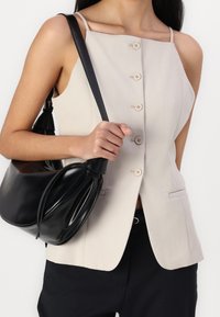 Top sin mangas beige con escote cuadrado, cierre frontal de botones y bolsillos laterales. Combinado con un bolso negro redondeado con detalle anudado.